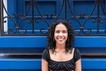 Young woman with curly hair smiling while standing in front of a vibrant blue wall with decorative ironwork.